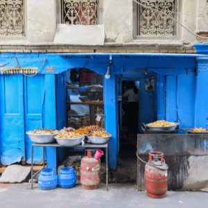 A blue store front kitchen in Nepal with two tables outside of it. Food in serving platters is spread across the tables. 