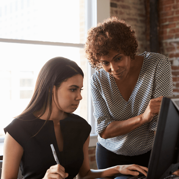 An older woman is standing over a young woman who is working at a computer at a desk. They are in front of windows. The older woman is pointing out something on the computer screen, and the young woman is taking notes.