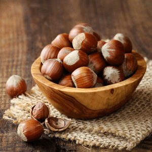A wooden bowl is full of hazel nuts. The bowl sits on a folded up piece of burlap on top of a wooden surface. 