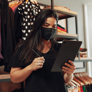 A Latina Entrepreneur is in a shop and holding a tablet and stylus while wearing a face mask.