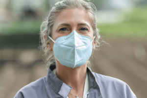 A blonde woman stands in an agriculture field with a tractor out of focus behind her. She is wearing a light blue KN95 face mask and a gray jacket.