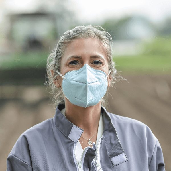 A blonde woman stands in an agriculture field with a tractor out of focus behind her. She is wearing a light blue KN95 face mask and a gray jacket.