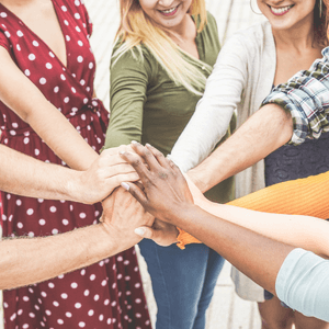 Nine women are standing in a circle and each of them has an arm outstretched into the center of the circle, and each has their hands on-top of the others'.