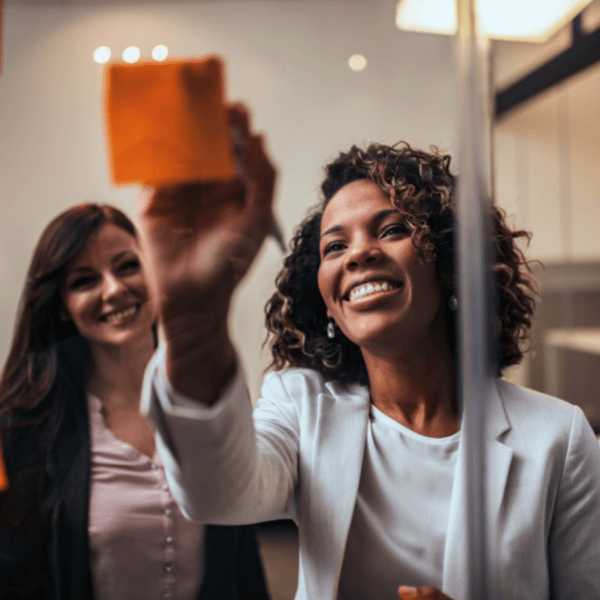 Two women are standing next to each other looking at a Post-It Note. One woman is writing on the orange Post It Note that is posted on a glass wall in their office.