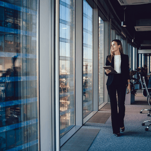 A woman in a pant suit is walking along the hall of a high rise office building. She is next to a wall of windows. 