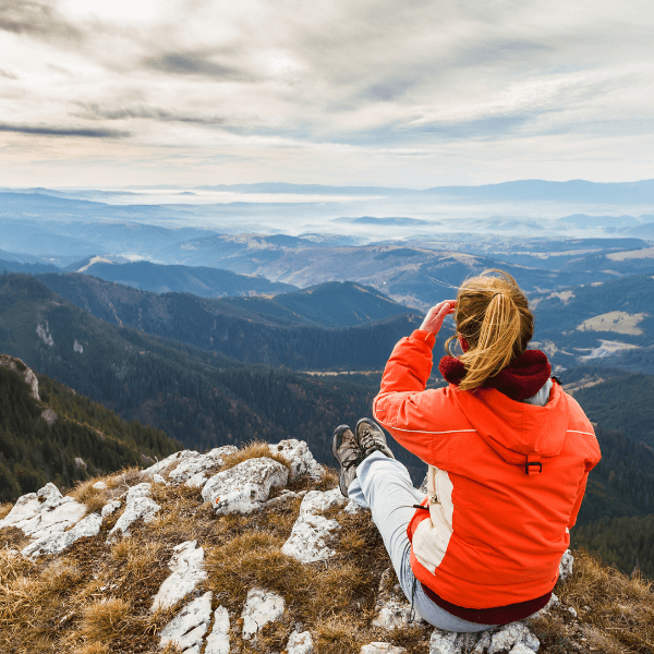 A woman in an orange raincoat is sitting on a rock with her back to the camera. She is overlooking a mountain range.