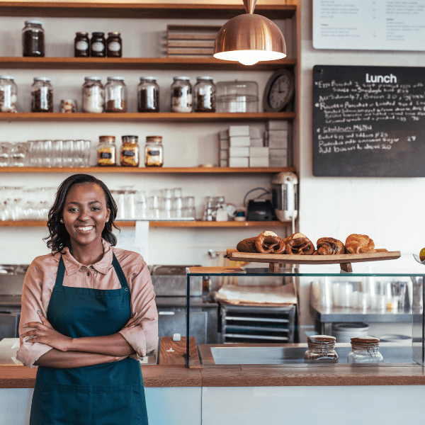 A woman wearing a blue apron stands in front of the counter of a cafe/bakery.