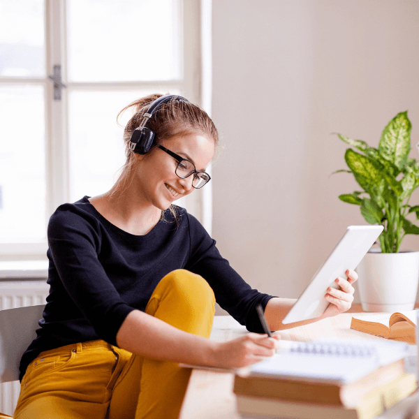 A woman sitting at a desk and wearing headphones. She is looking at a tablet while taking notes.