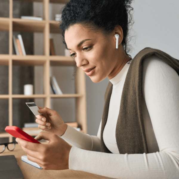 A woman is sitting at a desk holding a smartphone and a credit card. She has headphones in and is in front of a book shelf.