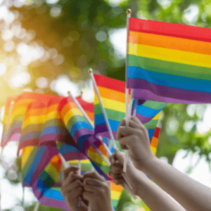 The hands of three different people holding up handheld rainbow pride flags that are on sticks with trees in the background.