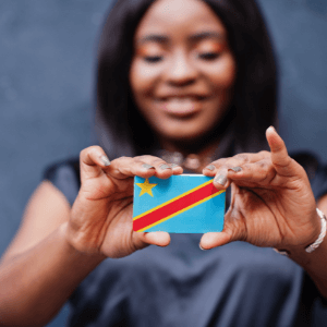 A congolese woman is standing in front of a blue wall and holding a small Democratic Republic of Congo flag in her hands. Her eyes are closed and she is wearing a navy blue shirt. 