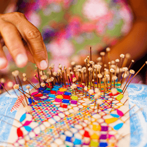 A woman's hand is shown taking a pin out of a multi-colored patchwork pin cushion.