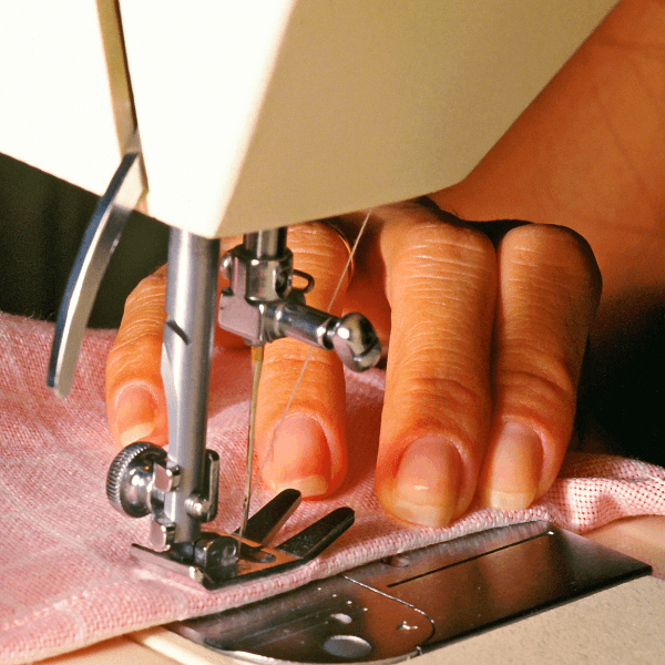 A close up photo of a woman's hand while she is using a white sewing machine to make a stitch in a piece of pink fabric.