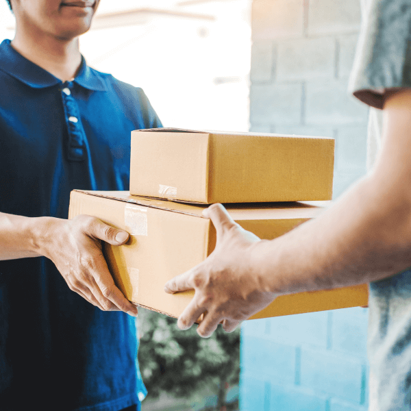 Two men face each other as one man hands the other a stack of two packages outside of his home.