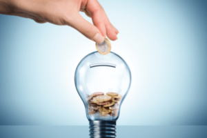 A hand places a coin into an unlit light bulb that is acting as a piggy bank. The image is against a light blue background.