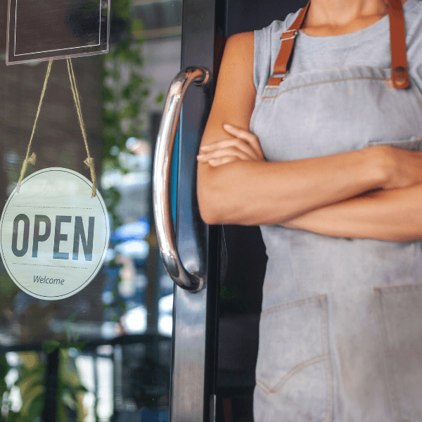 A small business owner stands outside of their store front. Their arms are crossed, and they are wearing a grey apron around their neck. Their head is outside of the shot. There is an "Open" sign on the door next to them.