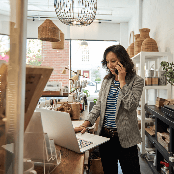 A woman stands in her shop while talking on her cellphone and looking at a computer.