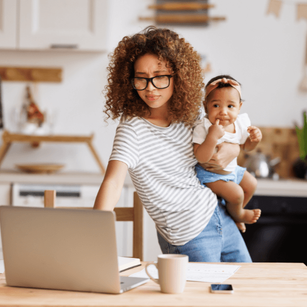 A woman standing and holding a baby on her hip as she is looking at a laptop computer screen.