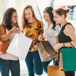 Four women stand next to each other while holding shopping bags. They are looking into their bags and laughing. 
