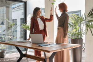 Two women in pantsuits are standing in an office space behind a desk and high-fiving each other.