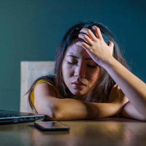 A woman sitting in a desk chair and leaning onto her desk stares sadly at her phone, with a hand on her forehead in frustration.
