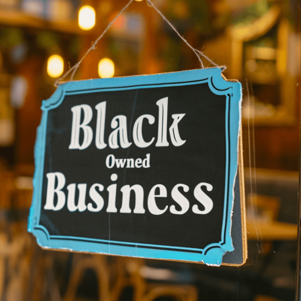 A black sign with an aqua blue border reads "Black Owned Business" hanging in a storefront window.