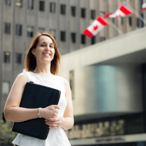 A woman standing outside of a Canadian embassy building with two Canadian flags on poles flying.