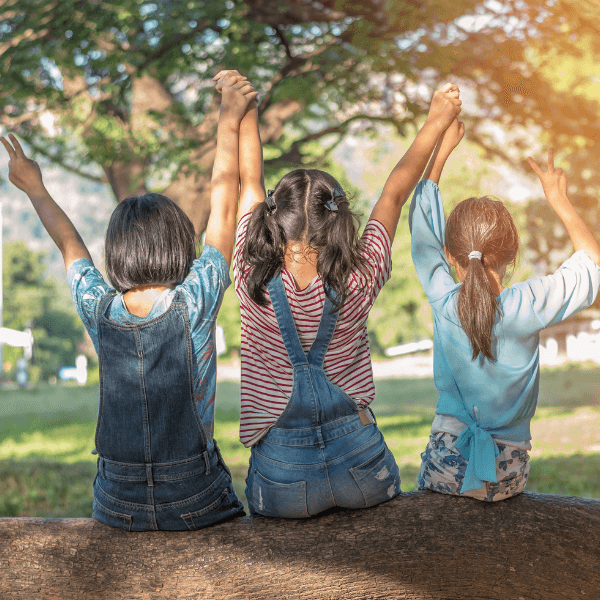 Three girls sitting on a tree branch with their arms in the air and holding hands.