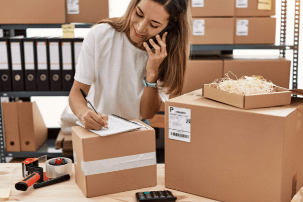 A woman on the phone and taking notes amidst shipping boxes.