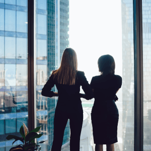 Two women in pantsuits staring out of a window looking at a skyscraper.