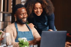 An African-American man sits at a desk using a laptop while an African-American woman stands next to him and leans over his shoulder looking at the laptop with him.