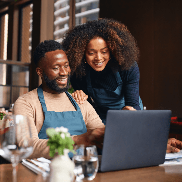 An African-American man sits at a desk using a laptop while an African-American woman stands next to him and leans over his shoulder looking at the laptop with him.