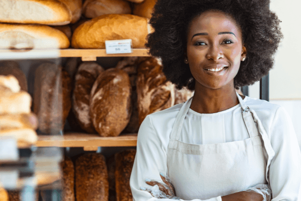 An African-American woman stands in front of shelves of loaves of bread. She is wearing a white apron, with her arms crossed and smiling at the camera.