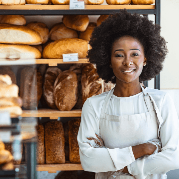 An African-American woman stands in front of shelves of loaves of bread. She is wearing a white apron, with her arms crossed and smiling at the camera.